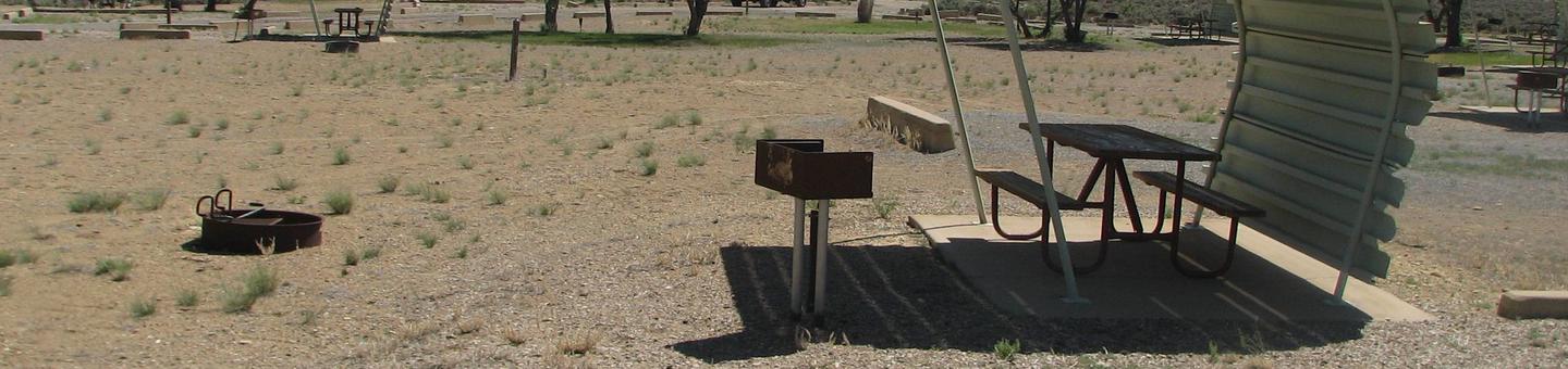 This site has a partially covered picnic table on a slab of cement with a grill next to it held up by a metal rod in the ground and a fire pit found in the gravel with very little grass.Buckboard Crossing Campground: Loop B, Site 27