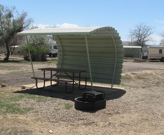 This site has a partially covered picnic table on a slab of cement and a fire pit with a grill grate found in the gravel. Another site can be seen in the background.Buckboard Crossing Campground: Loop A, Site 29