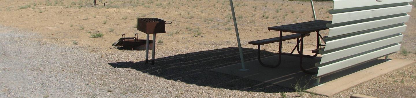 This site has a partially covered picnic table on a slab of cement with a grill next to it held up by metal rods in the ground and a fire pit with a grill grate found in the gravel.Buckboard Crossing Campground: Loop B, Site 29