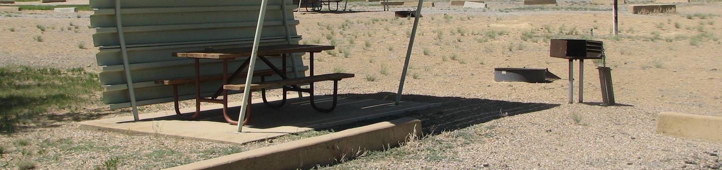 This site has a partially covered picnic table on a slab of cement with a grill next to it held up by a metal rod in the ground and a fire pit found in the gravel.Buckboard Crossing Campground: Loop B, Site 3