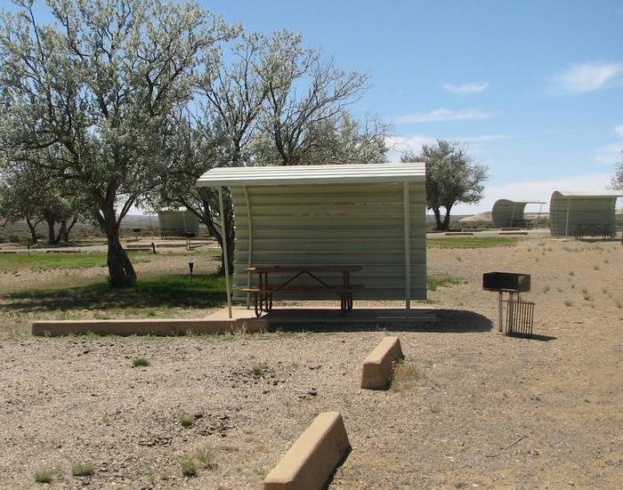 This site has a partially covered picnic table on a slab of cement with a grill next to it held up by a metal rod in the ground and a fire pit found in the gravel.Buckboard Crossing Campground: Loop B, Site 3