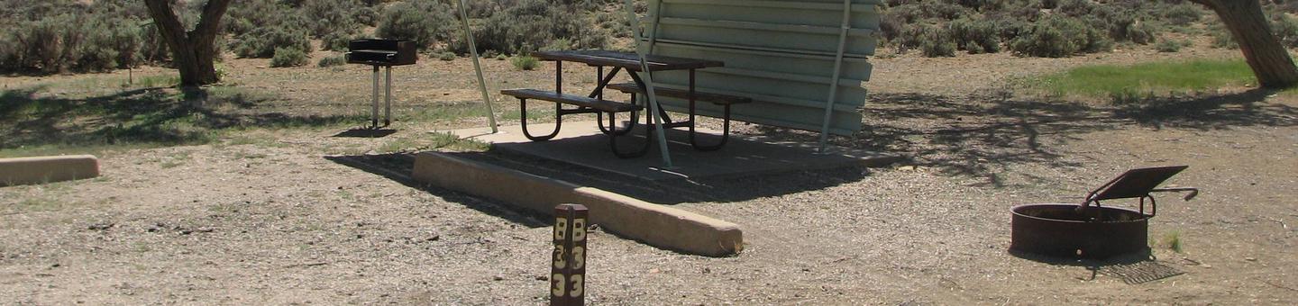 This site has a partially covered picnic table on a slab of cement with a grill next to it held up by a metal rod in the ground and a fire pit found in the gravel.Buckboard Crossing Campground: Loop B, Site 33