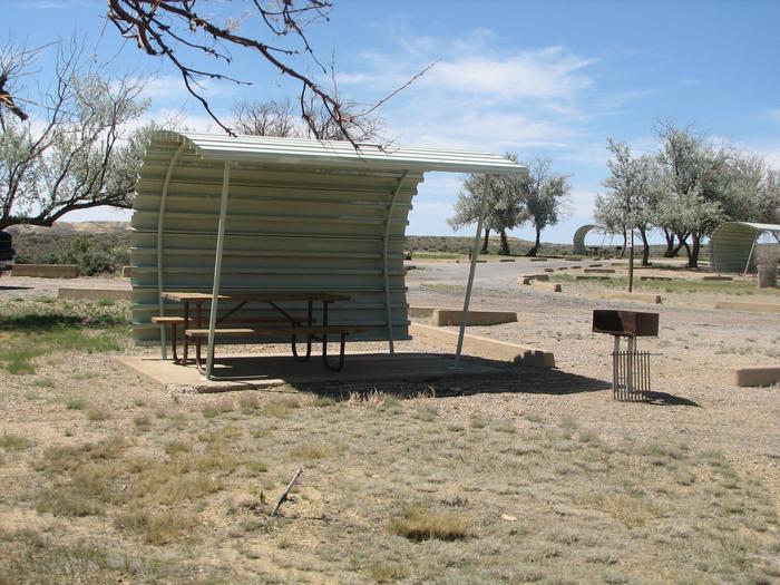 This site has a partially covered picnic table on a slab of cement with a grill next to it held up by a metal rod in the ground. Some shade can be found in this site. Other camp sites can be seen in the background.Buckboard Crossing Campground: Loop B, Site 34
