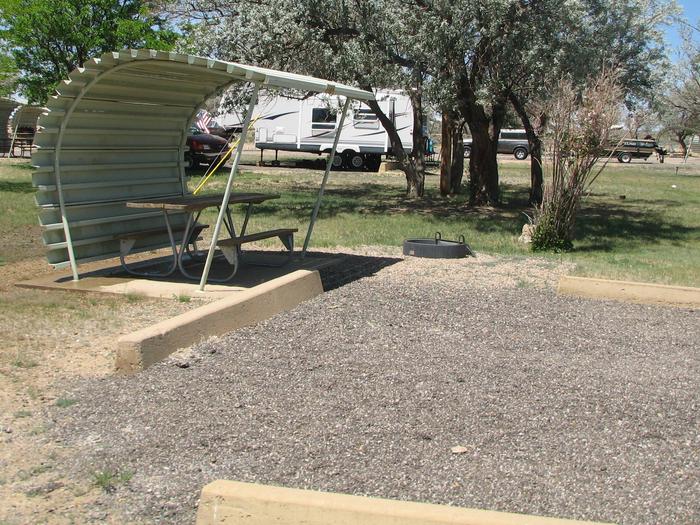 This site has a partially covered picnic table on a slab of cement and a fire pit found in the gravel. There is some shade and grass in this site.Buckboard Crossing Campground: Loop A, Site 5