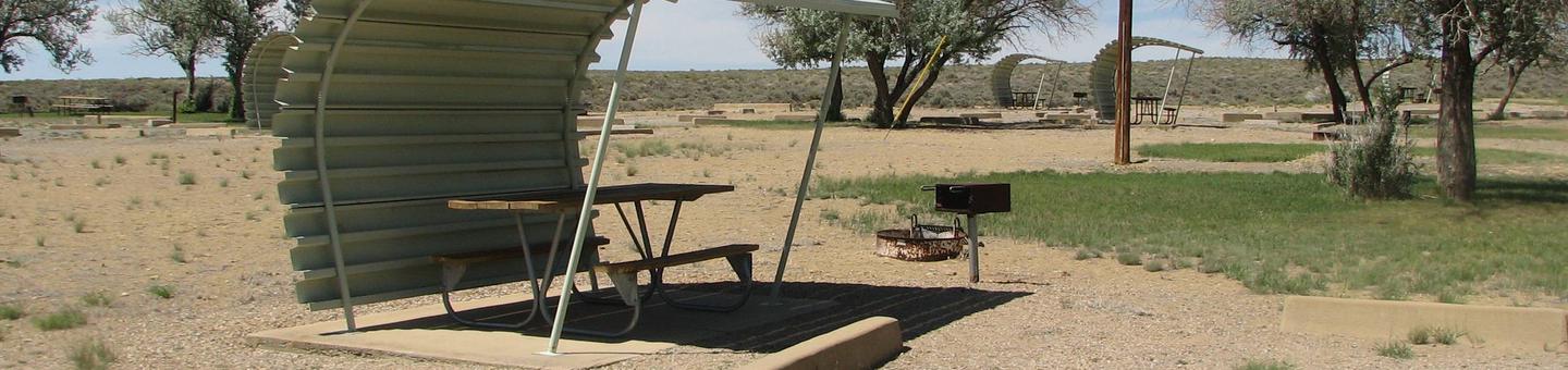 This site has a partially covered picnic table on a slab of cement with a grill next to it held up by a metal rod in the ground and a fire pit found in the gravel.Buckboard Crossing Campground: Loop B, Site 5
