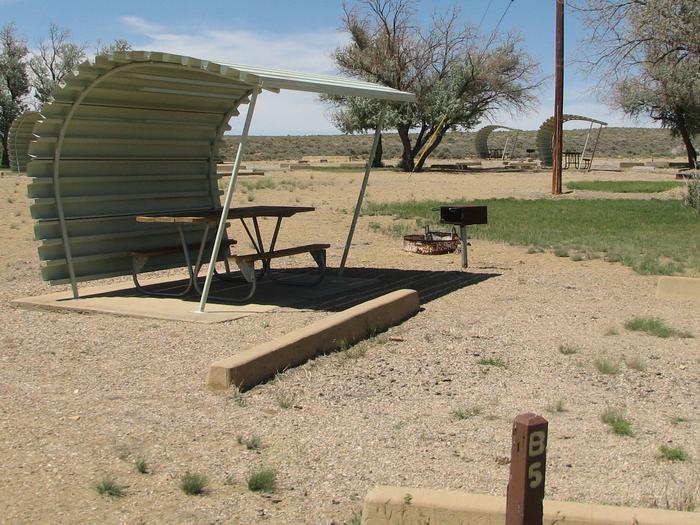 This site has a partially covered picnic table on a slab of cement with a grill next to it held up by a metal rod in the ground and a fire pit found in the gravel.Buckboard Crossing Campground: Loop B, Site 5