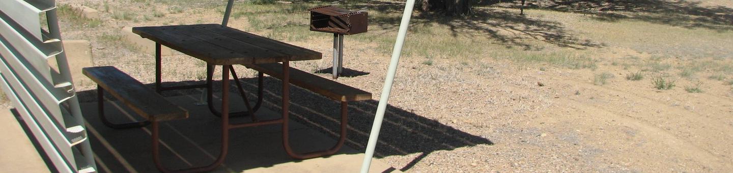 This site has a partially covered picnic table on a slab of cement with a grill next to it held up by a metal rod in the ground.Buckboard Crossing Campground: Loop B, Site 6