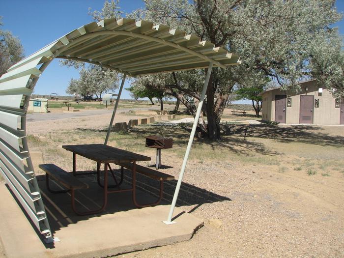 This site has a partially covered picnic table on a slab of cement with a grill next to it held up by a metal rod in the ground.  Tree with shade and the restroom facilities are close by.Buckboard Crossing Campground: Loop B, Site 6