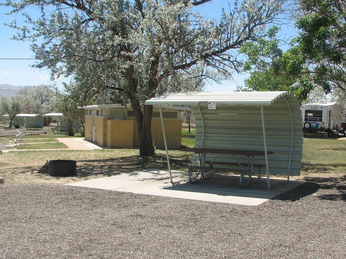 This site has a partially covered picnic table on a slab of cement and a fire pit found in the gravel. Shade trees, grass and restroom facilities are nearby.Buckboard Crossing Campground: Loop A, Site 8