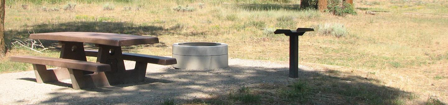 This site has a picnic table and a fire pit in a gravel area. Trees and grasses surround the area.Canyon Rim Campground: Site 7