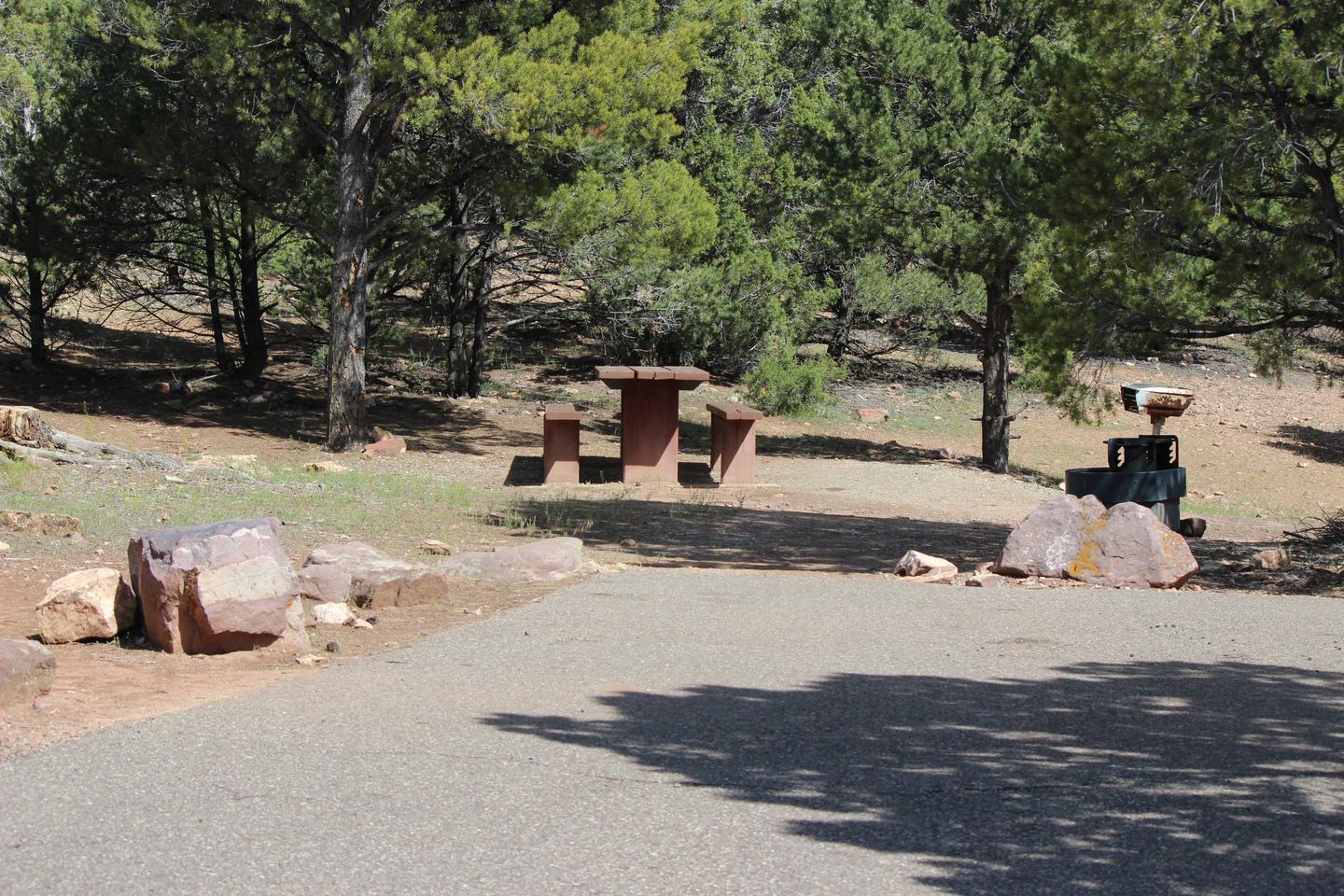 This site has a picnic table and fire pit with a grill grate in a gravel area behind a parking area. There are a number of trees in the background.Cedar Springs Campground: Site 3
