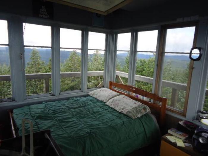 Bed in front of window filled corner with view of mountains and forest outside.1Clear Lake Cabin Lookout sleeping area.