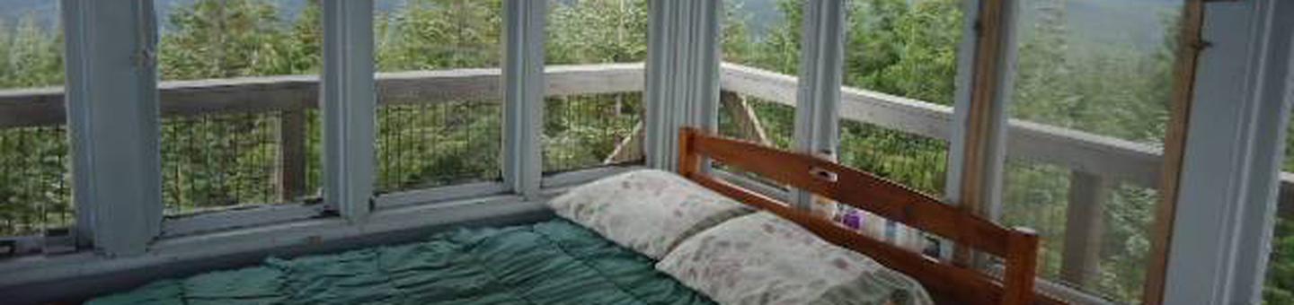 Bed in front of window filled corner with view of mountains and forest outside.Clear Lake Cabin Lookout sleeping area.