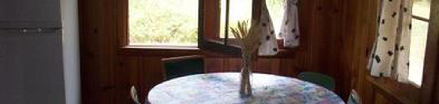Refrigerator next to round table covered with floral cloth, set with dried flower arrangement, circled by four chairs looking through open window and white patterned curtains to sunny, forested yard.TANEUM CABIN