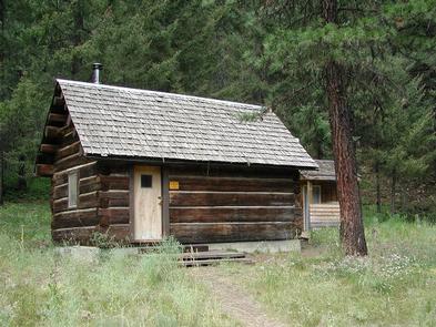 Small log cabin next to pine tree in open glade in front of conifer forest.TEANAWAY GUARD STATION