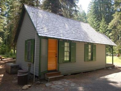 Gray cabin with three windows with green shutters and two steps up to  one wood door in front of a small lawn and conifer forest.BOX CANYON GUARD STATION CABIN
