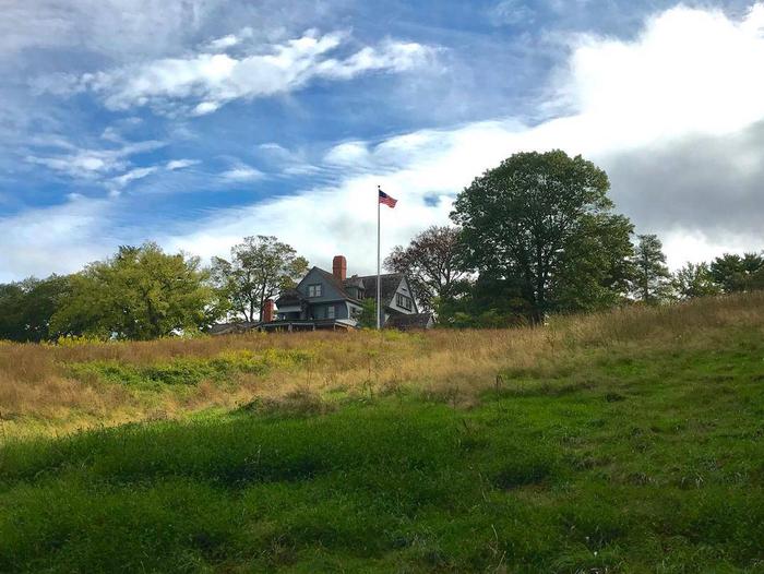 A gray house a top a grassy hill with blue sky and clouds in the background.Theodore Roosevelt's home atop Sagamore Hill.