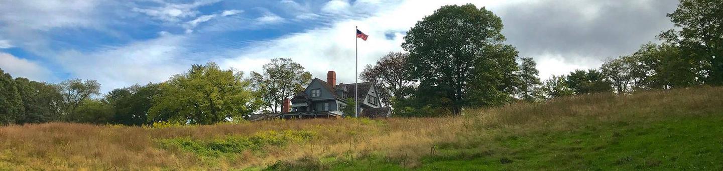 A gray house a top a grassy hill with blue sky and clouds in the background.Theodore Roosevelt's home atop Sagamore Hill.