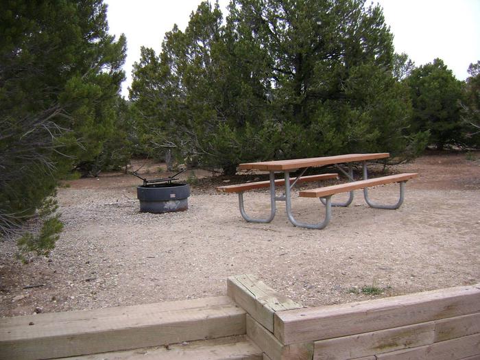 Picnic table and fire pit in a gravel area with trees surrounding the area. There are steps and wood wall that separate the parking and picnic areas.Deer Run Campground: Site 1