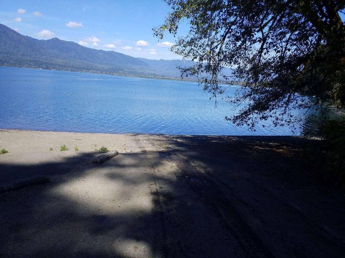 Shaded boat ramp leading to blue, sunlit lake with mountains and clouds floating in blue sky as backdrop.Willaby Campground