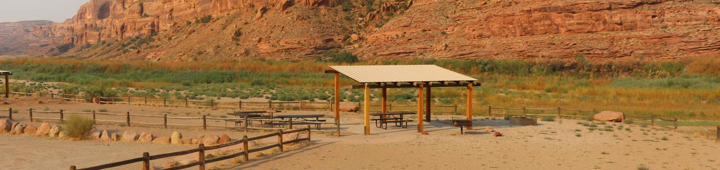 Gold Bar Group Site B shade shelter, picnic tables, and parking and tent area near the Colorado River with tall red rock cliffs in the distance.