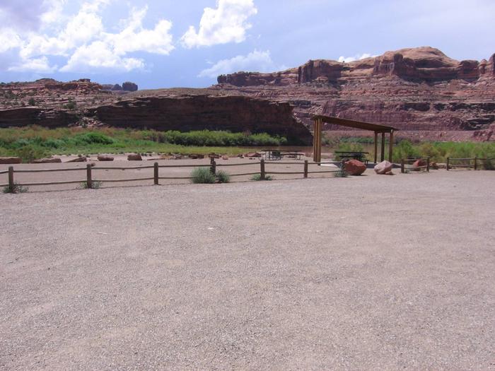 Shade shelters, picnic tables, parking area, and tent area at Gold Bar Group Site A. In the near distance is the Colorado River and rounded, red rock cliffs.