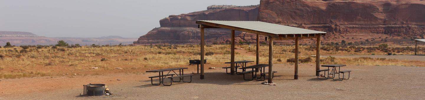 Lone Mesa Group Site C shade shelter, picnic tables, and fire ring. Large, red rock, sandstone walls in the distance.