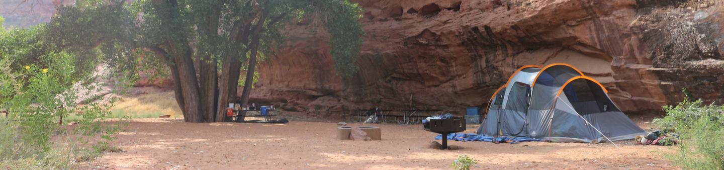 A tent is set up on a large, open, sandy surface next to a red rock canyon wall in the shade. There is also a standing grill next to the tent.