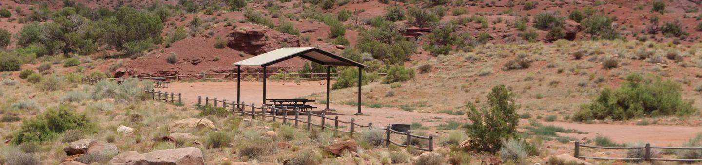 The Upper Onion Creek Group Site B has a large shade shelter with picnic tables underneath and a fire ring nearby. Surrounded by undulating land covered in small shrubs and juniper trees.