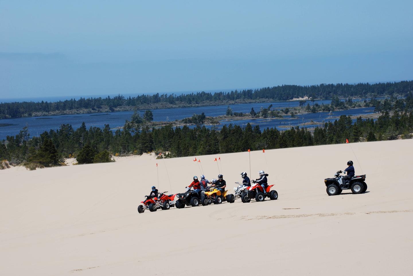 Off road vehicles on flat expanse of sand in front of blue lake ringed by conifer trees.The dunes near Horsfall Campground