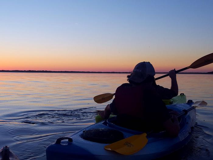 A young man paddles a kayak across still water at sunset.