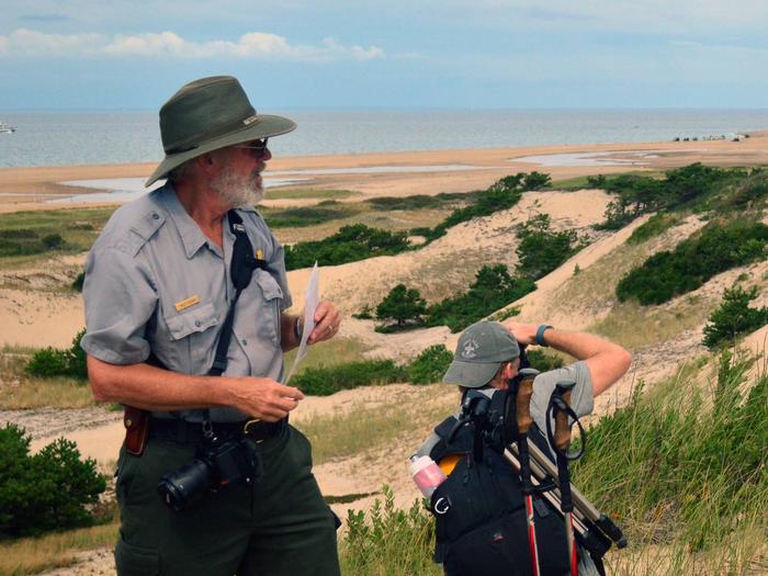 A ranger looks on from atop a sand dune as a man takes a photograph of the landscape.