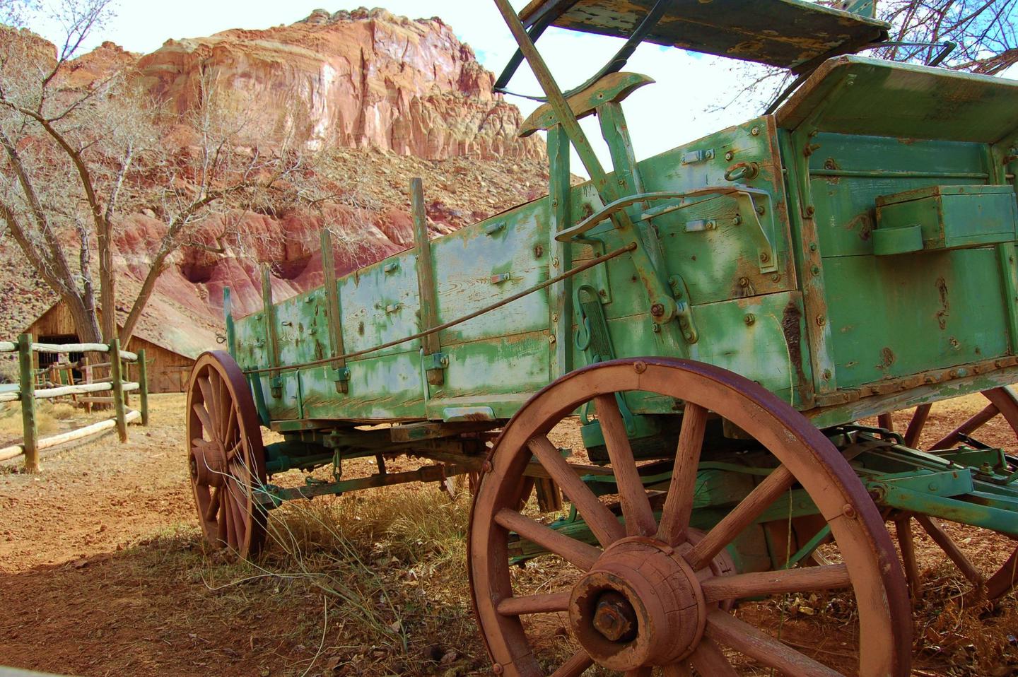 A green wagon with rusty wheels is the center of the image. A red rock cliff is behind it.Old wagon near Fruita Campground