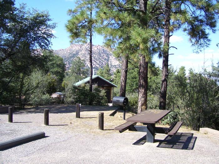 Yavapai Campground Site 6 shares parking with Site 7. Table, ballards separating parking, camping and grill area with toilet and Granite Mountain in background. Shade over tent area. Yavapai Campground Site #6