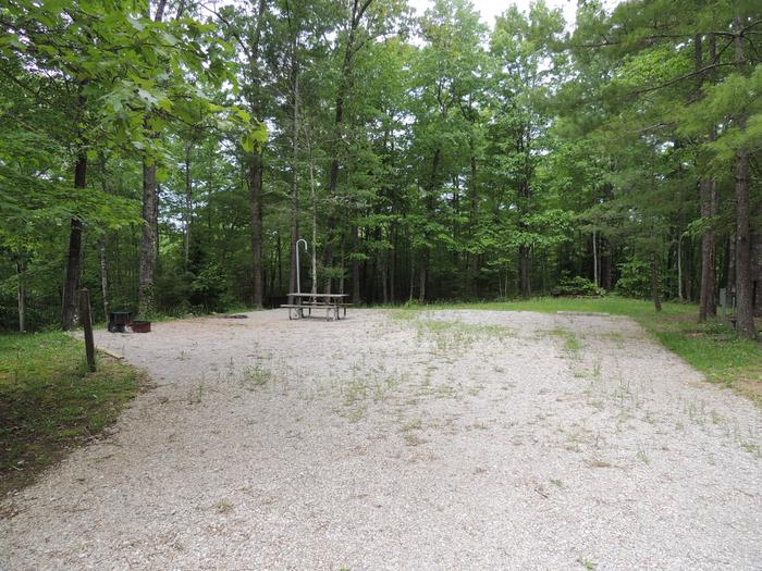 Gravel parking alongside tent pad with picnic table and lantern hook. Green trees surround the site.Site 12