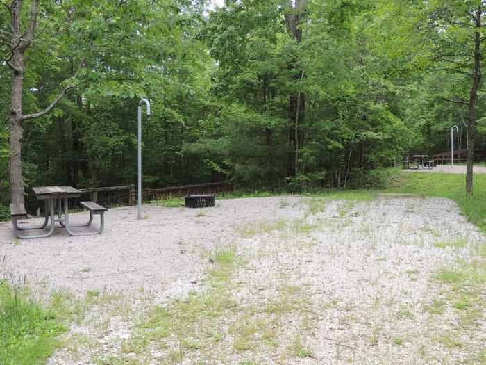 Gravel parking alongside tent pad with picnic table and lantern hook. Green trees surround the site.Site 14