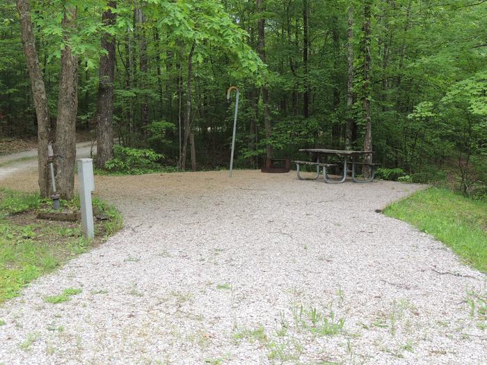 Gravel parking alongside tent pad with picnic table and lantern hook. Green trees surround the site.Site 15