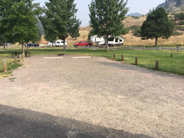 Site 4 at Holter Lake Campground. Graveled campsite with picnic table and fire pit in the background. Graveled site surrounded by wooden posts to keep parking limited to the graveled and paved surfaces. 3 large trees in the background.Site 4 BLM Holter Lake Campground.