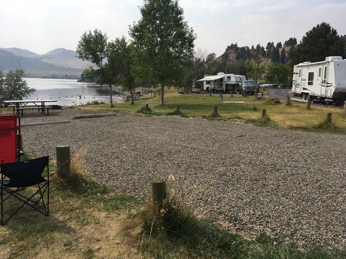 Site 5 at Holter Lake Campground. Graveled campsite with wooden posts and paved access. Picnic table and fire pit in the background. View of Holter Lake Campground. Trees dispersed throughout campground.Site 5 Holter Lake Campground.