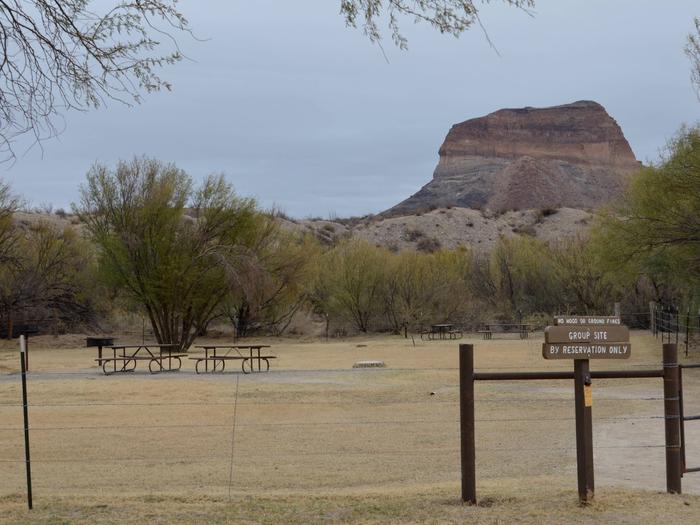 Group campsite sits among the desert brush in a fenced in area with a desert bluff in the backgroundCampsite and picnic tables sit among the desert brush in a fenced in area