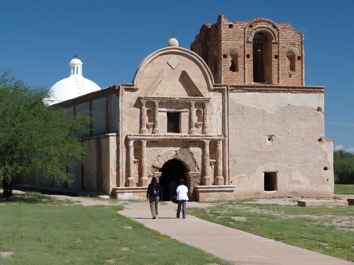 Park visitors walking on a trail towards the ruins of the Franciscan church at Mission San José de Tumacácori.The Tumacácori mission church was built in the early 1800's and is maintained in a state of arrested ruin.  The goal is to preserve the original structure rather than additional restoration or speculative completion of architectural features.