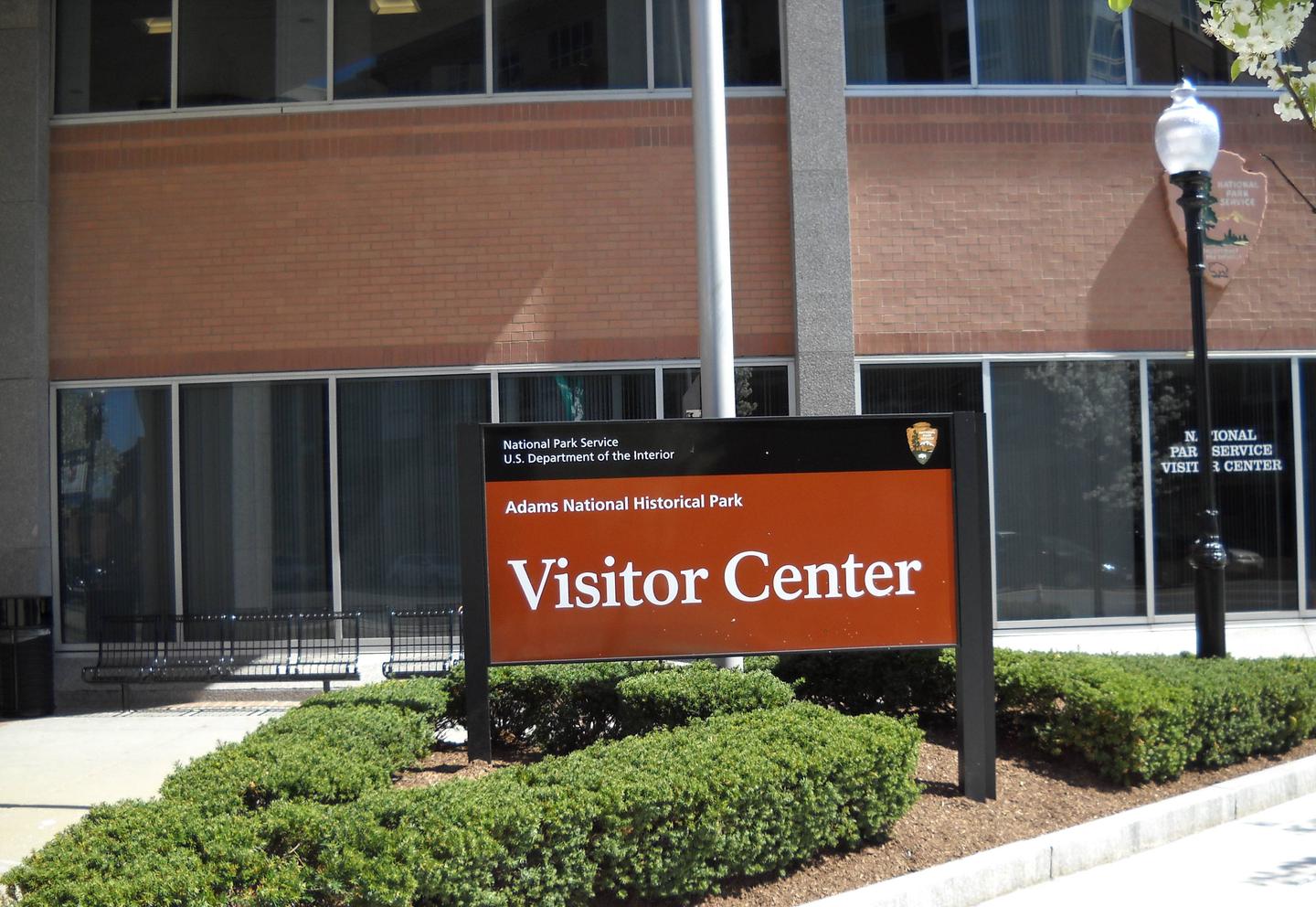 A dark orange sign with white text reads "Adams National Historical Park - Visitor Center." Planted around the sign are small green shrubs. There is a modern building with tall windows and a tall black lamp post behind the sign. The Adams National Historical Park Visitor Center Sign