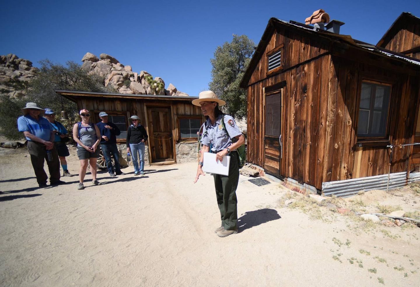 A woman wearing a NPS uniform is addressing a group of people in front of a historic ranch.Tours at Keys Ranch give visitors a unique experience to learn about the Keys family and their survival in a harsh desert environment.