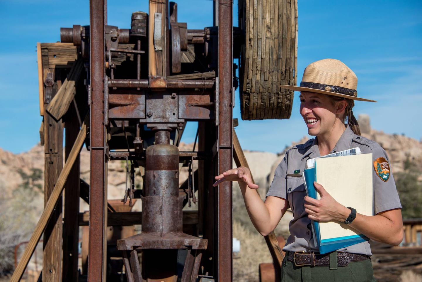 A woman wearing a NPS uniform is standing in front of an old gold mill.Gold mining was a way of life for many settlers in the region and old gold mills can still be found. 