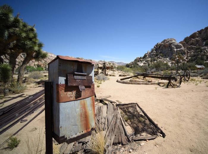 A rusted tin box stands on a post with the words "Keys mail" scratched into the side. In the background there is various rusted ranch equipment.Mail call! How long do you suppose it would take for a letter to arrive at Keys Ranch?
