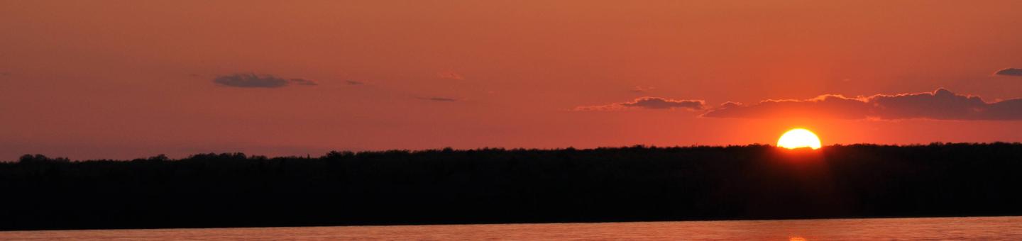The sun setting, silhouettes a forested island, creating an orange sky, which is reflected in the foreground lake water. Sunset at Apostle Islands National Lakeshore