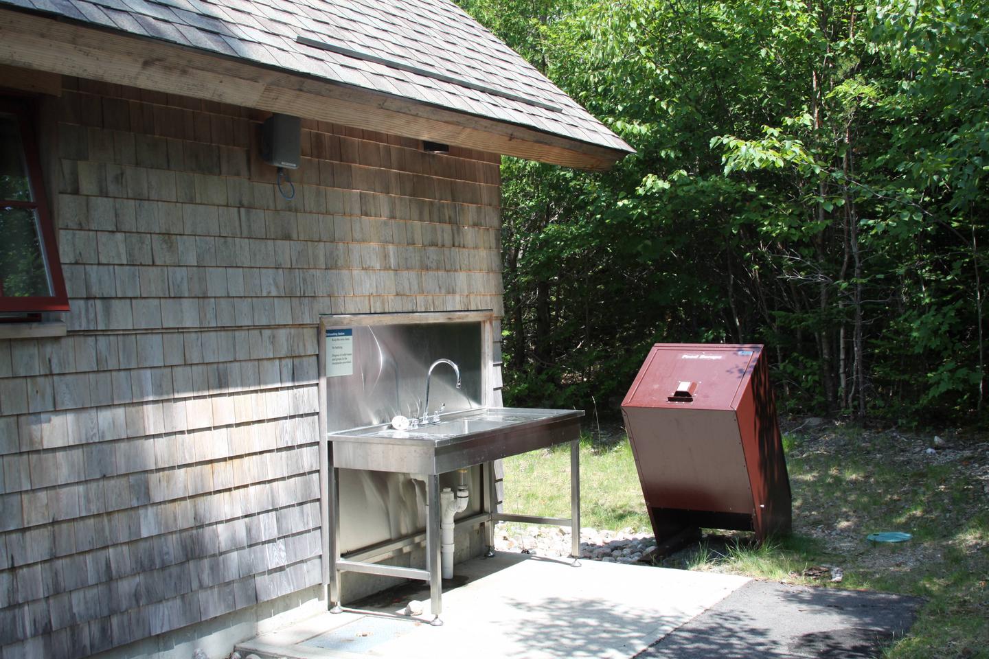 Stainless steel sink on outside of a cedar shingle sided building with bear proof trash can.A typical dish-washing Station