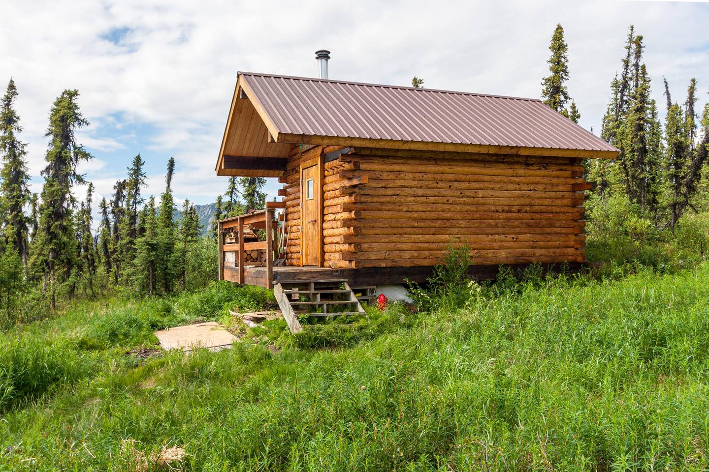 A cabin sits on a hillside surrounded by green grass and spruce trees.Borealis-LeFevre Cabin in summer