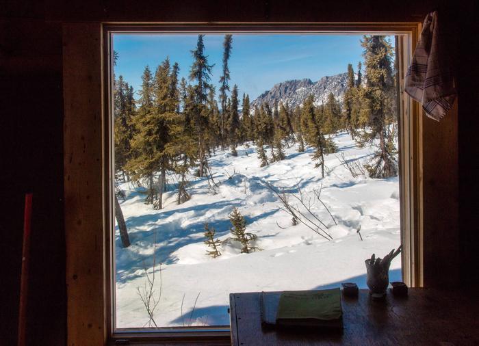 View through a window shows a snow-covered forest and a distant, rocky ridge.Winter view from Borealis-LeFevre Cabin.