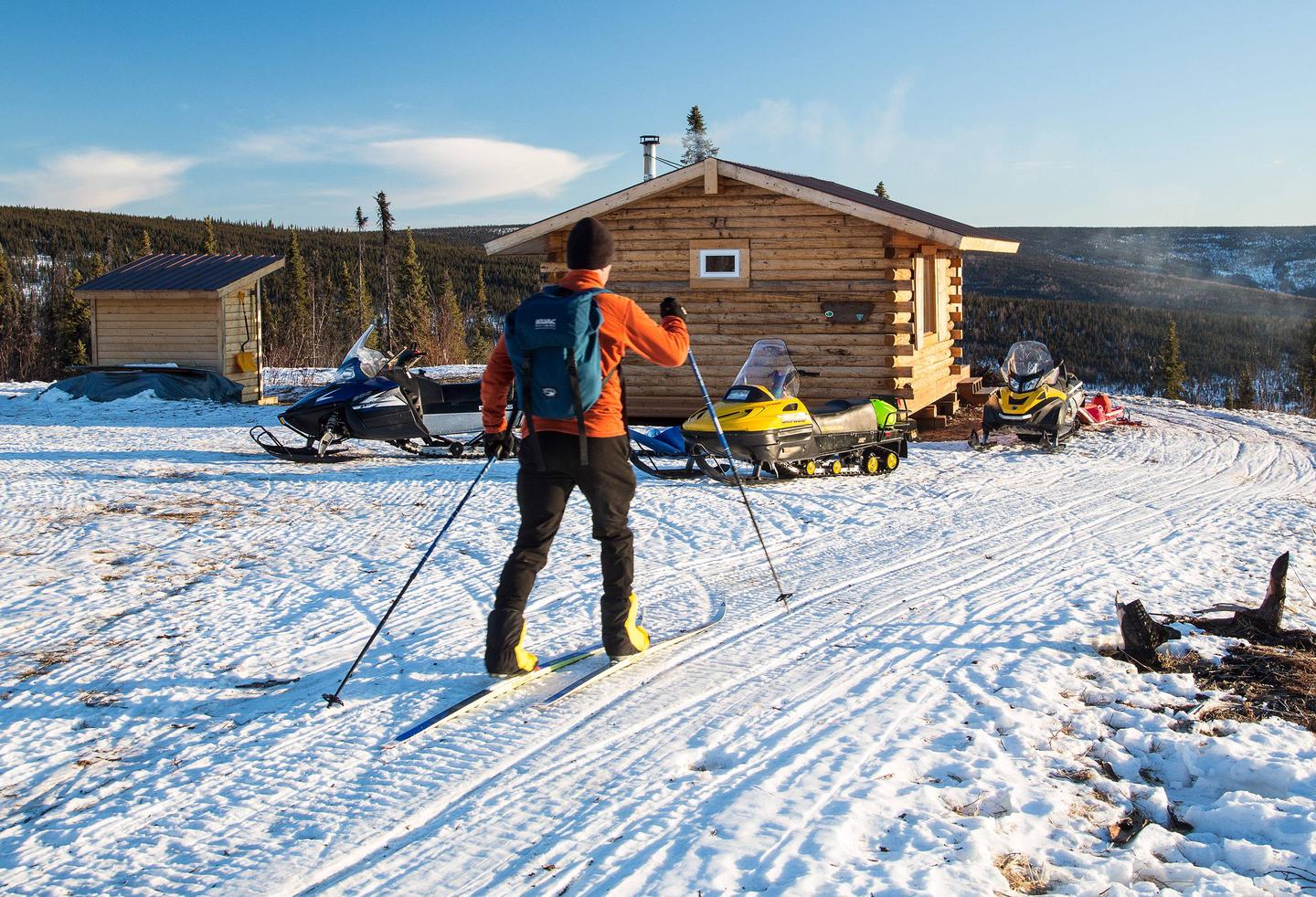 A man skies up to a log cabin surrounded by snowmobiles.Arriving at Moose Creek Cabin on skis.